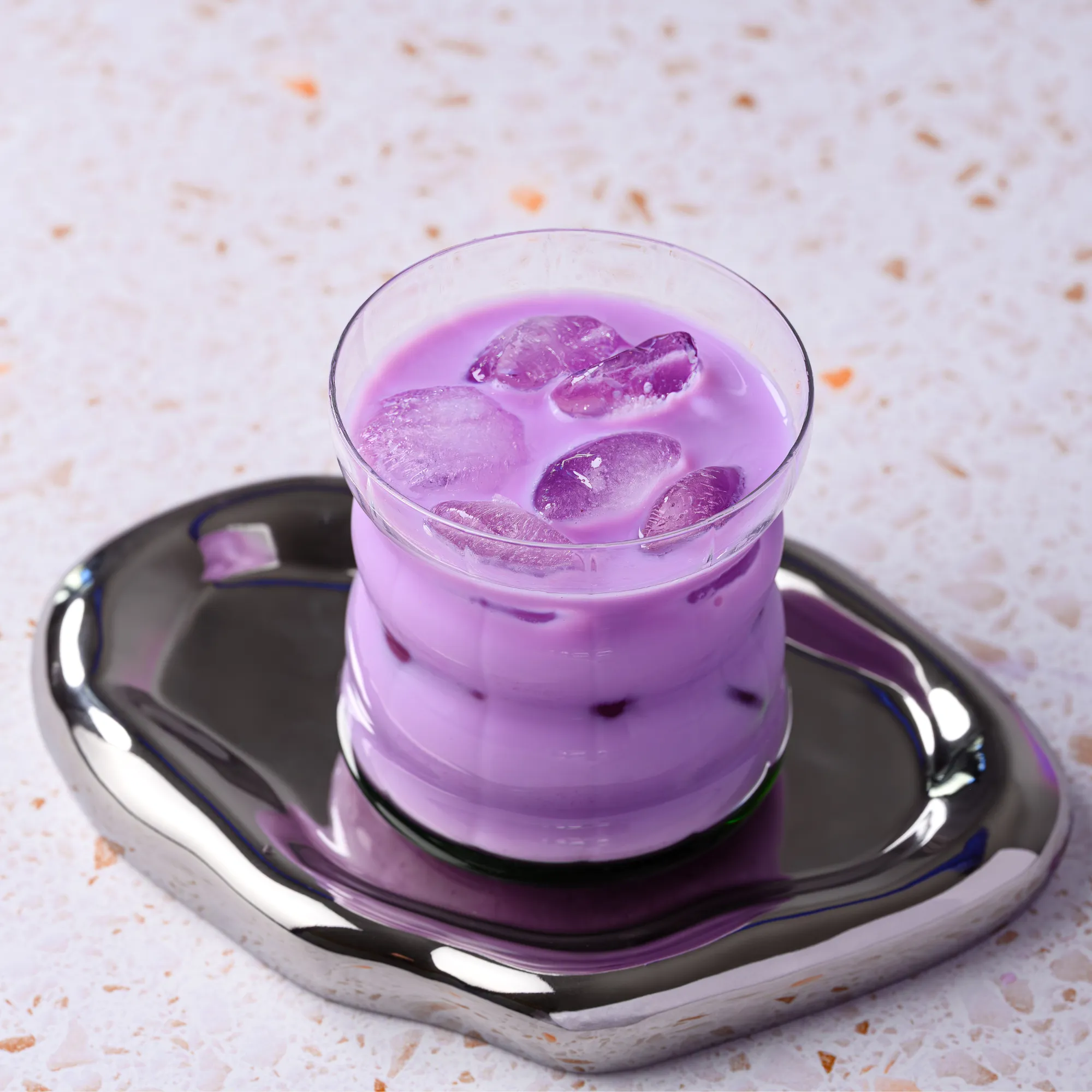 Ube latte glacé mauve, avec des glaçons, dans un verre posé sur un plateau argenté.